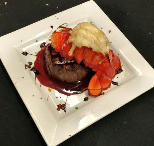 A gourmet dish featuring a grilled filet mignon topped with lobster, tomatoes, and a rich sauce on a white plate.