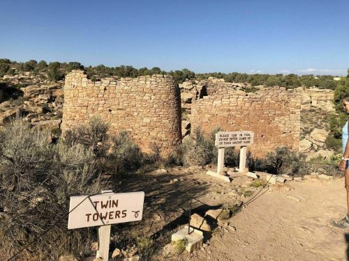 Two stone ruins labeled "Twin Towers" with a trail sign nearby, surrounded by desert vegetation and rocky terrain.