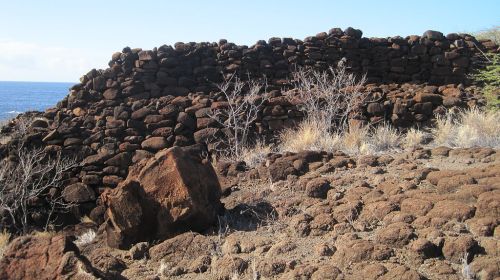 Ancient stone wall ruins by the ocean, surrounded by dry vegetation and rocky terrain under a clear blue sky.