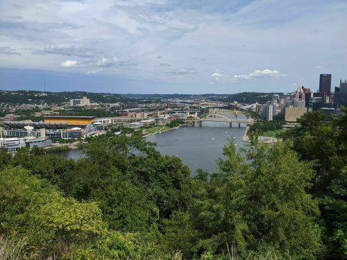 A panoramic view of Pittsburgh, featuring rivers, bridges, and city skyline under a partly cloudy sky.
