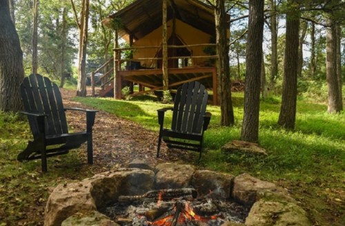 A cozy campsite with two chairs by a fire pit, surrounded by trees and a cabin in the background.