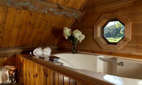 Cozy wooden bathroom with a large tub, flowers in a vase, and an octagonal window overlooking greenery.