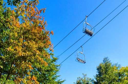 Two empty ski lift chairs suspended above colorful autumn trees under a clear blue sky.