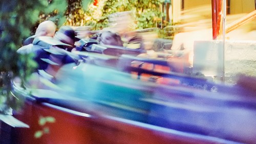 A blurred image of people in motion, possibly on a ride, surrounded by greenery and colorful lights.