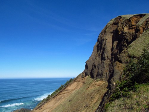 A rugged cliffside overlooking a calm ocean under a clear blue sky, with greenery in the foreground.