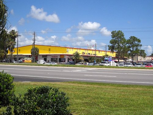 A bright yellow building with shops, surrounded by palm trees and a clear blue sky, viewed from across a road.
