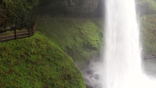 A lush green landscape with a waterfall cascading down, surrounded by rocky cliffs and a wooden railing path.