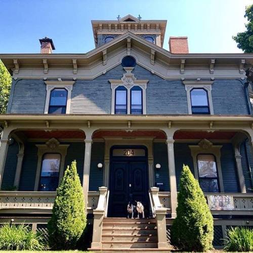 A large, historic blue Victorian house with a black door, flanked by two green shrubs and two dogs on the steps.