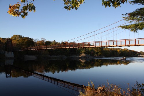 A suspension bridge spans a calm river, surrounded by autumn foliage and reflecting in the water below.