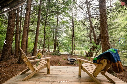 Two wooden chairs on a deck surrounded by tall trees, with a cozy blanket draped over one and a fire pit in the background.