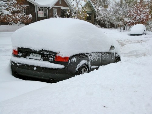 A car buried under a thick layer of snow, with snow-covered streets and houses in the background.