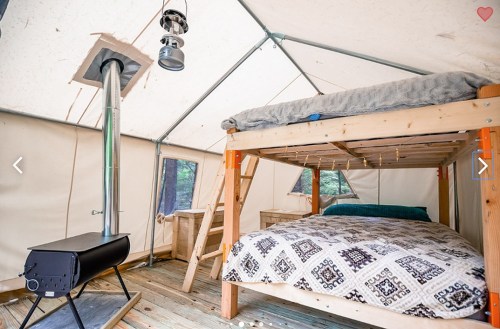 Cozy glamping tent interior with a bunk bed, wood stove, and warm lighting, surrounded by nature.