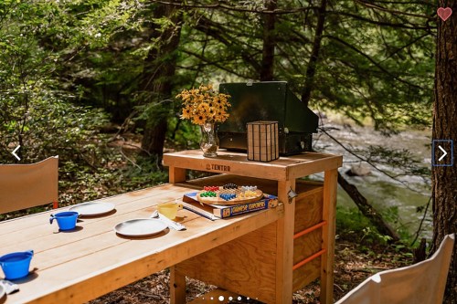 A wooden outdoor table set with plates, a game, and flowers, surrounded by trees near a flowing stream.