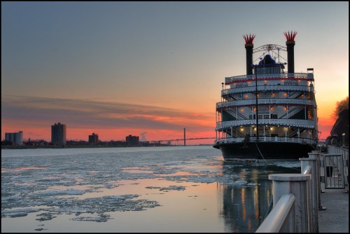 A riverboat with three smokestacks docked by the icy river at sunset, with city buildings in the background.