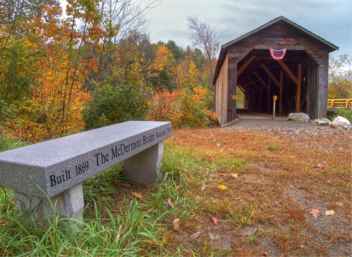 A stone bench with an inscription beside a rustic covered bridge, surrounded by autumn foliage.