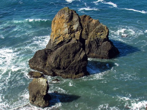 A large rock formation surrounded by waves in a blue ocean, with smaller rocks nearby.