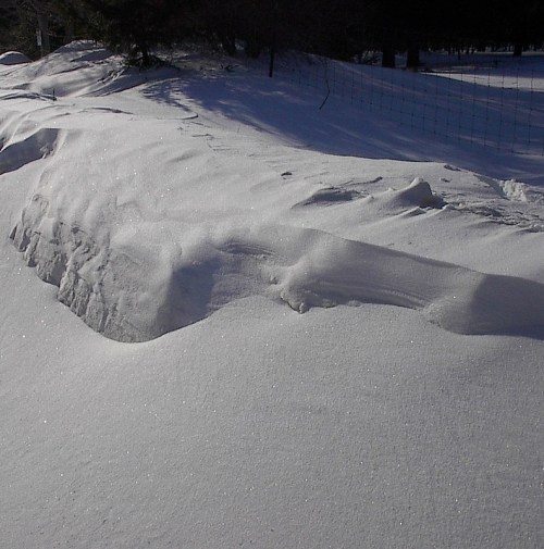 A snowy landscape with a smooth, undulating surface glistening in the sunlight, bordered by a fence in the background.