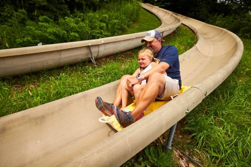 A man and a girl joyfully ride down a winding slide surrounded by greenery.