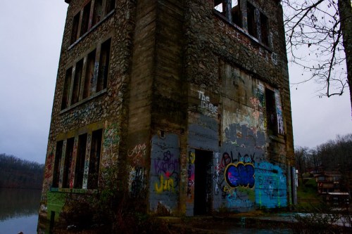 Abandoned stone building covered in graffiti, near a lake, surrounded by trees under a cloudy sky.