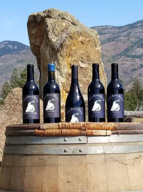 Five bottles of wine with labels displayed on a wooden barrel, set against a rocky backdrop and mountains.