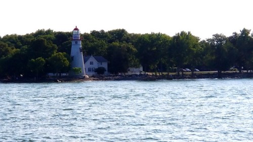 A lighthouse stands on a shore surrounded by trees, with calm water in the foreground and a clear sky above.