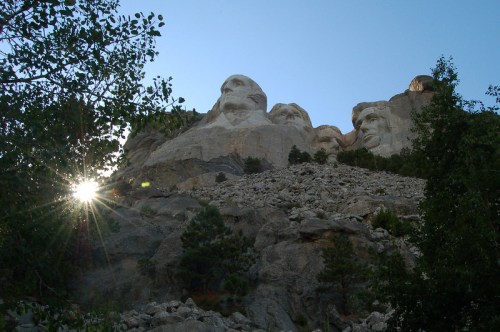 Mount Rushmore with four presidential faces carved into the rock, framed by trees and a setting sun in the background.
