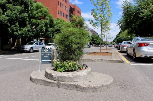 A small park with a sign, surrounded by greenery and flowers, located in a city street with parked cars nearby.