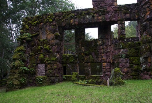 Ruins of a moss-covered stone structure surrounded by greenery and trees. Windows and doorways are partially visible.