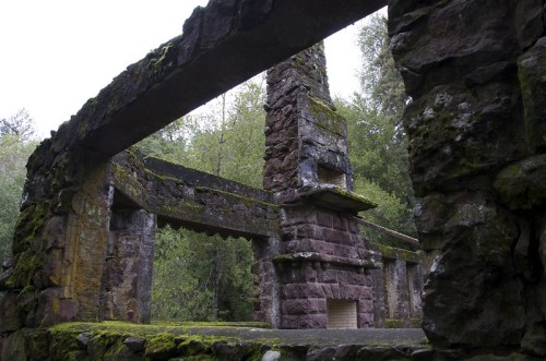 Ruins of a stone structure covered in moss, surrounded by trees and greenery, with a cloudy sky above.