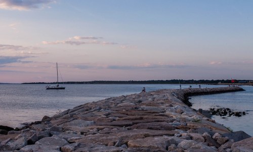 A rocky jetty extends into calm waters at sunset, with a sailboat nearby and people walking along the path.