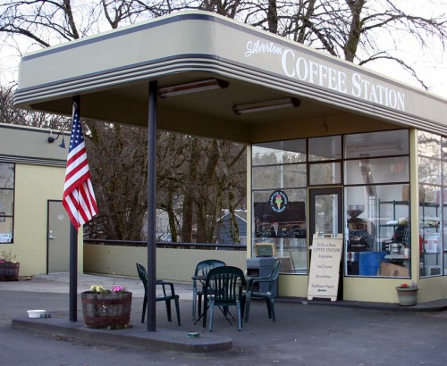 A coffee station with outdoor seating, an American flag, and a sign displaying menu items. Trees in the background.