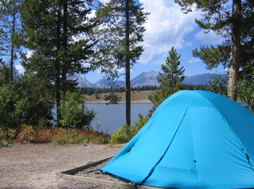 A bright blue tent by a serene lake, surrounded by trees and mountains under a partly cloudy sky.