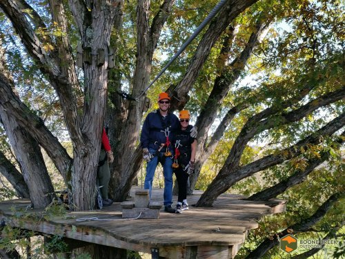 Two people in harnesses stand on a wooden platform among trees, smiling and ready for a zipline adventure.