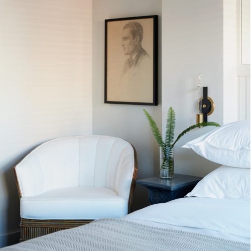 A cozy bedroom corner featuring a white chair, a small table with a fern, and a framed portrait on the wall.