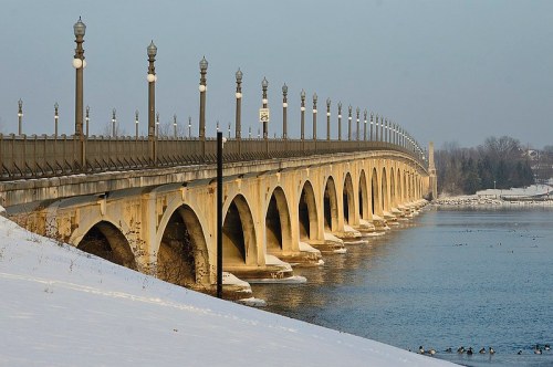 A long bridge with arches spans a river, lined with street lamps, surrounded by snow and calm water.