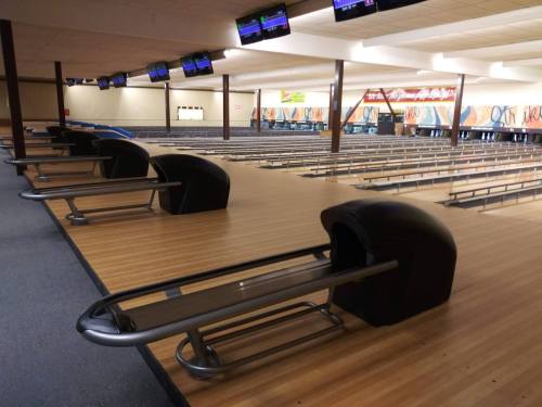 A spacious bowling alley with empty lanes and seating areas, featuring overhead score screens.