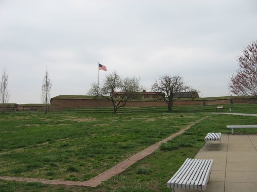 A grassy area with benches, featuring a fort in the background and an American flag flying above it.