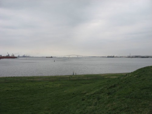 A calm body of water with a distant bridge, surrounded by green grass and a cloudy sky.