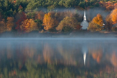 A serene lake reflects autumn foliage and a church steeple, shrouded in morning mist.
