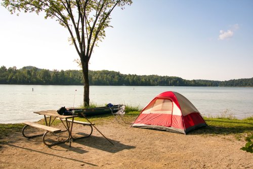 A red and gray tent by a calm lake, with a picnic table and a canoe nearby under a tree.