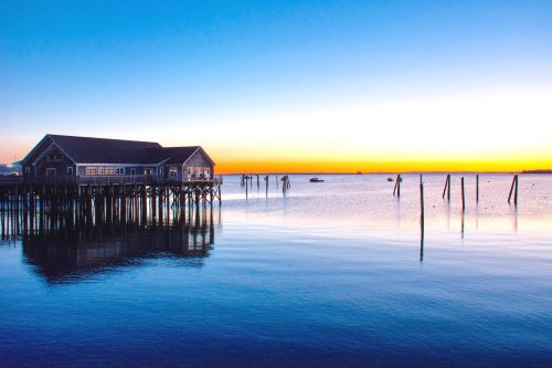 A serene sunset over calm waters, with a pier and a building reflecting on the surface.