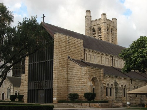 A stone church with a tall tower, large windows, and a cross on top, surrounded by trees and cloudy skies.