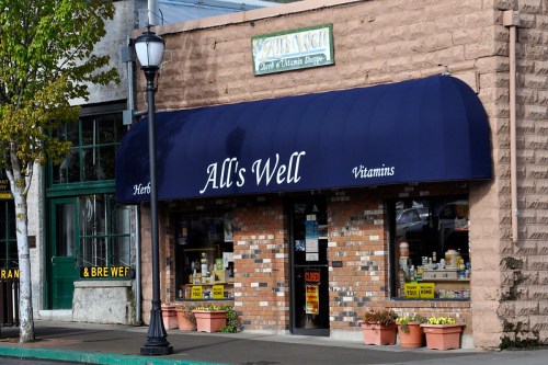 A storefront with a blue awning reading "All's Well" and displaying herbal and vitamin products in the window.