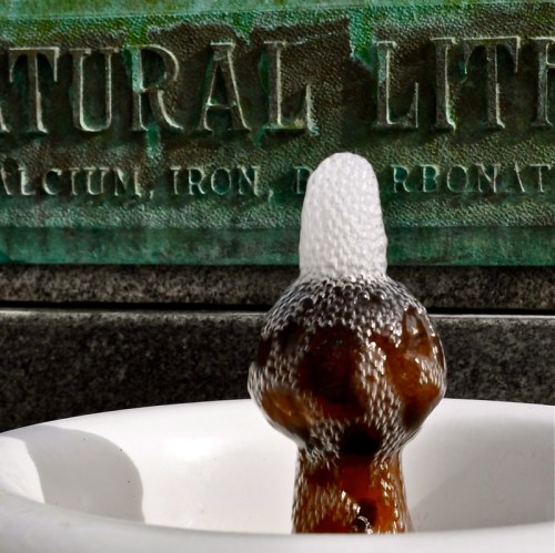 A close-up of a bubbling fountain with a textured stone background displaying the words "Natural Lithia."