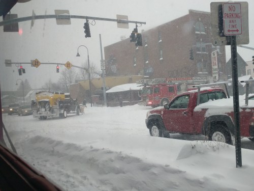 Snow-covered street scene with traffic lights, a fire truck, and a snowplow clearing the road in a winter storm.