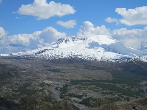 Snow-capped mountains under a blue sky with fluffy clouds, overlooking a valley with sparse vegetation.