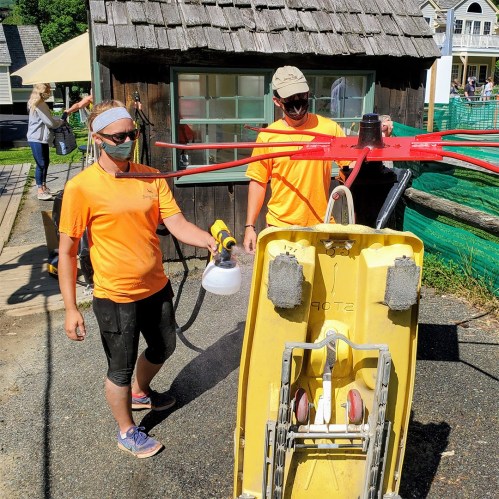 Two people in orange shirts are cleaning a yellow cart with a spray nozzle near a wooden building.