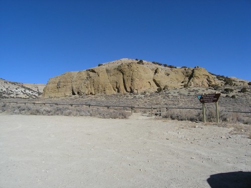 A rocky outcrop under a clear blue sky, with a signpost indicating a nearby trail or site. Dry, sparse vegetation surrounds.
