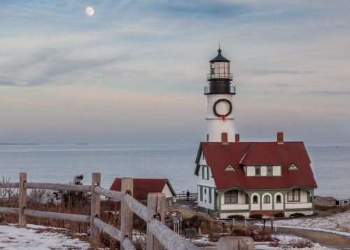 A lighthouse with a red-roofed house beside it, overlooking a calm sea under a full moon and cloudy sky.
