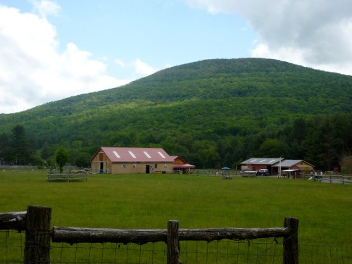 A scenic view of a farm with a barn, surrounded by lush green fields and a mountain in the background under a cloudy sky.